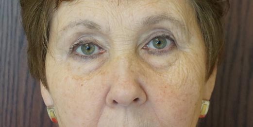 An older woman with short brown hair and green eyes, looking directly at the camera against a wood-panel background.
