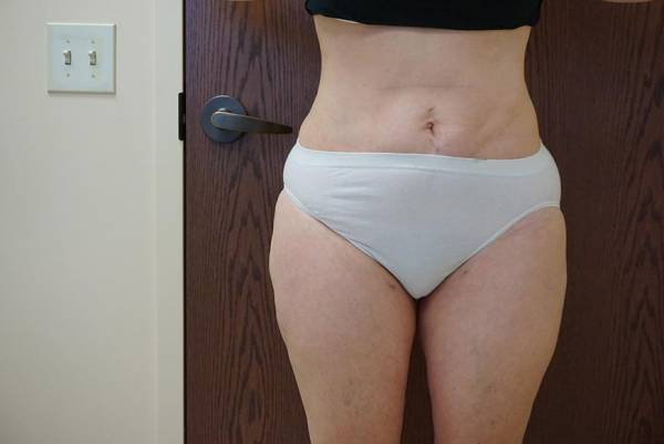 Person standing indoors in front of a closed wooden door, wearing a black top and white underwear, with midsection and upper legs visible.