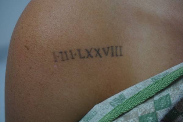 A tattoo on a person's shoulder displays the Roman numerals "III•LXXVIII".