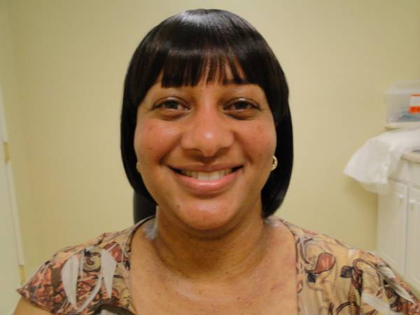 A woman with short black hair and bangs smiles at the camera. She is wearing a patterned blouse and gold hoop earrings, seated indoors against a light-colored wall.