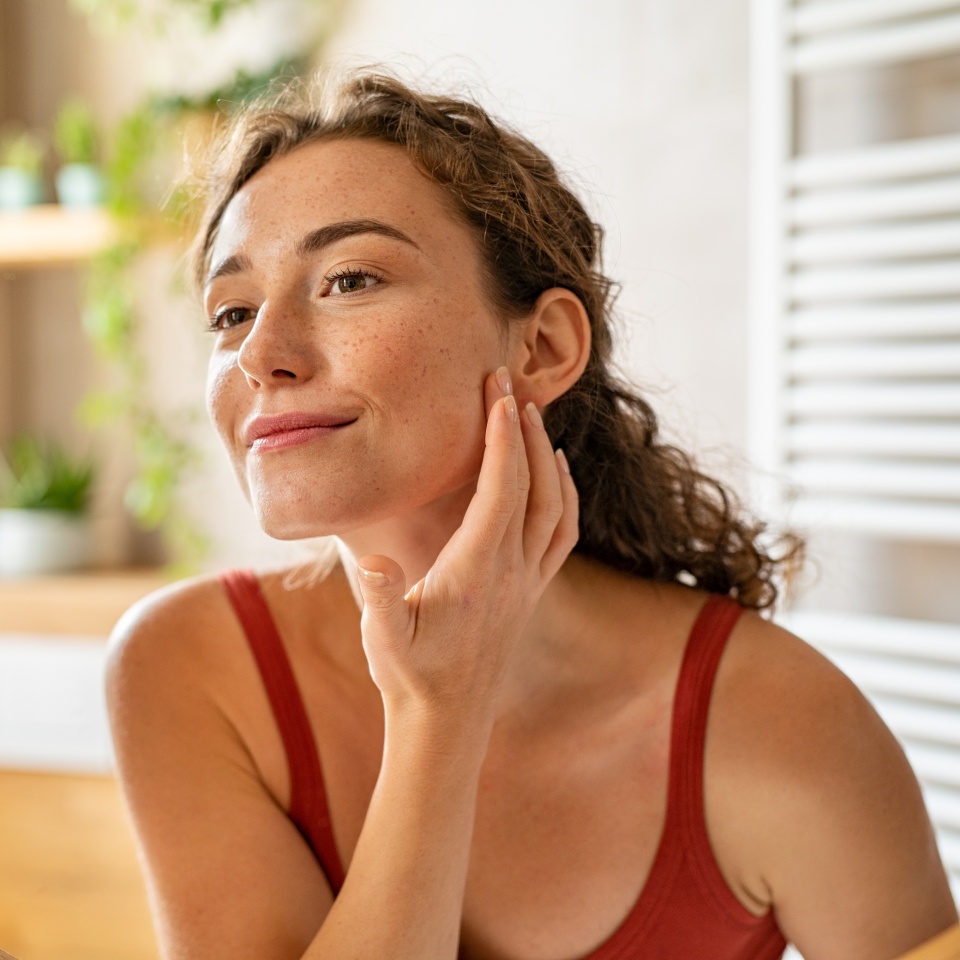 Beauty girl looking at mirror while touching her face and checking pimple, wrinkles and bags under the eyes, during morning beauty routine. Happy smiling beautiful young woman applying moisturizer.