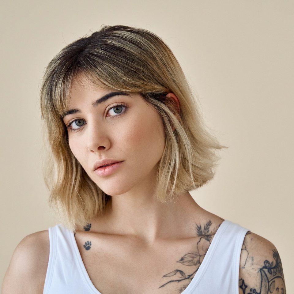 Cool blonde pretty gen z girl female stylish model short blonde hair beautiful face healthy skin and tattoos looking at camera wearing white top isolated at beige background. Portrait. Copy space