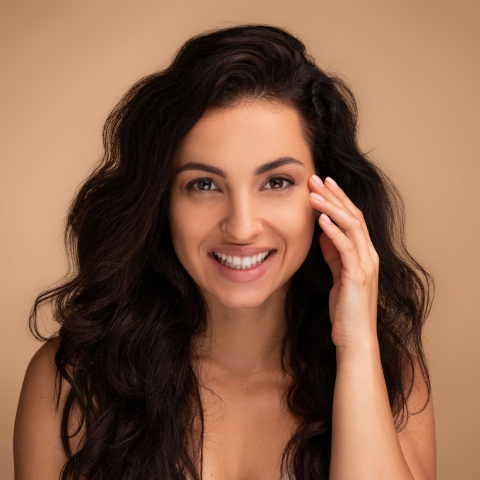 Anti-aging eye care treatment, aesthetic procedures concept. Attractive brunette long-haired young woman touching under eyes skin and smiling, closeup, beige studio background