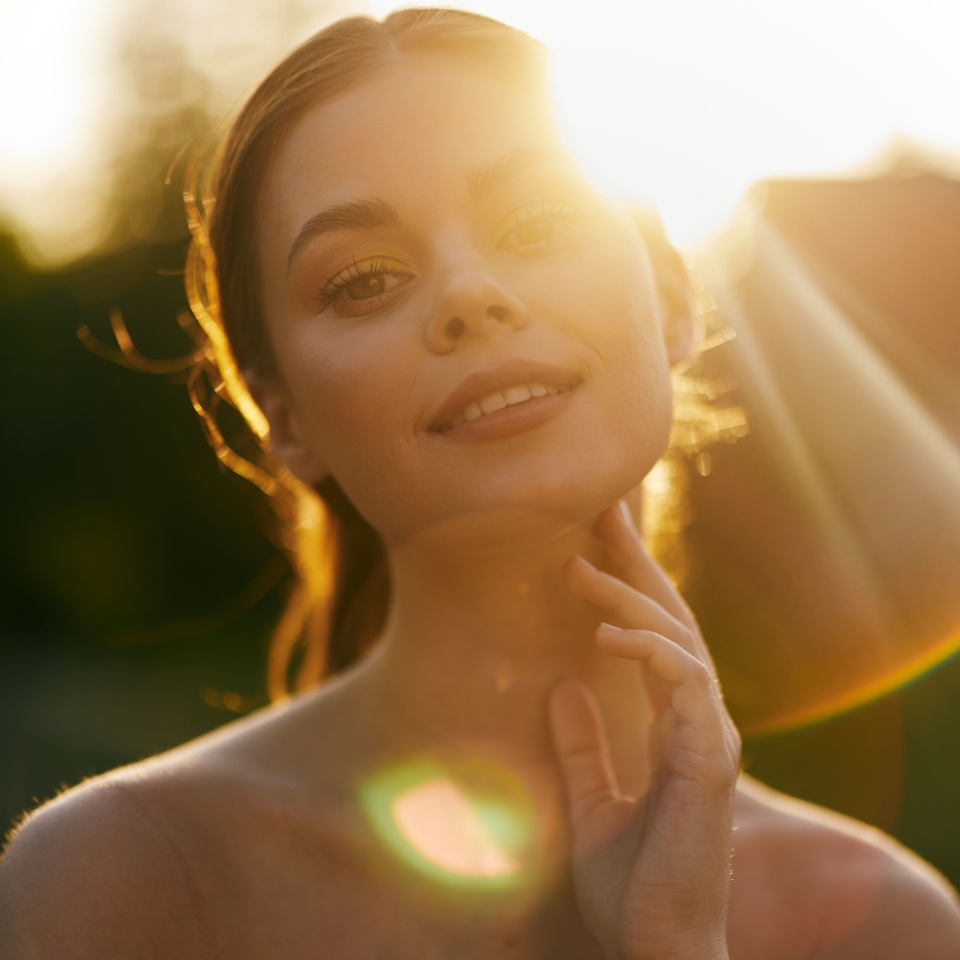 Young woman with glowing skin smiles serenely in natural sunlight, evoking warmth and beauty, surrounded by greenery and sunshine