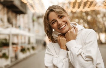 A young woman in a white hoodie smiles and poses outdoors on a city street with string lights in the background.