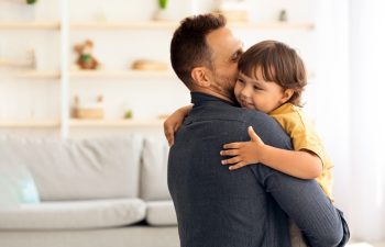 Cute little boy enjoying time with loving daddy, caring man embracing and kissing his lovely son, holding him on hands at home interior