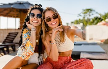stylish smiling beautiful women relaxing having fun on beach in summer fashion trend outfit, colorful apparel boho style, tropical vacation wearing sunglasses, using smartphone taking selfie picture